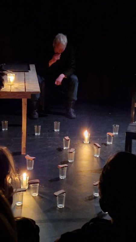 Сцена уставлена поминальными стаканами вокруг зажжённой свечи; актёр сидит у стола, создавая траурную и ритуальную атмосферу финала.