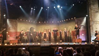 Финальная сцена постановки «Фигаро» с ансамблем актёров на фоне больших условных декораций и зрителями в переднем ряду.