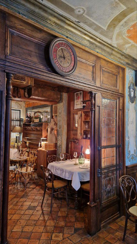 Уютный обеденный уголок ресторана с деревянной мебелью, круглым столом и оформлением в ретро-стиле, видна декоративная дверь и часы над ней.