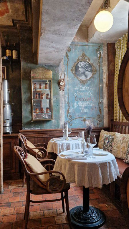 Интерьерный ракурс с небольшим столиком у окна, белая скатерть, мягкие диванные сиденья и расписные стеновые панно в стиле старой французской аптеки.