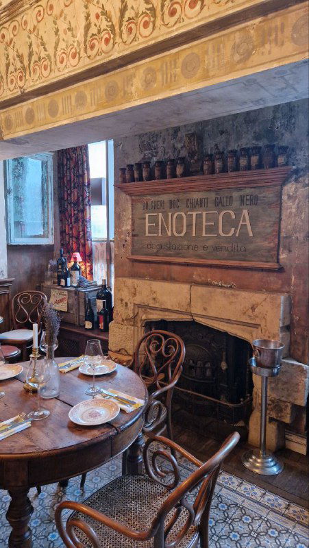 Общий вид зала с камином и деревянной мебелью, столики накрыты скатертями, декоративные надписи и винные ящики создают атмосферу энотекы.