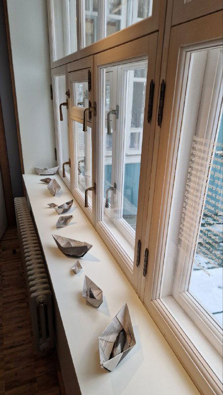 Подоконник с небольшими бумажными корабликами и документами — тонкая инсталляция, связывающая личные истории и материальность экспозиции.