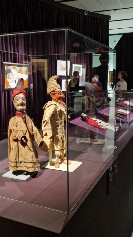 Стеклянная витрина с куклами, масками и предметами декоративного искусства на выставке «Под маской» в Музее русского импрессионизма.
