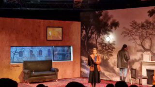 Интерьер декорации: диван и фон с живописным панно, актёр у дивана создает камерную домашнюю атмосферу сцены.