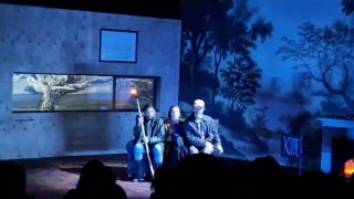 Три актёра сидят на скамье в приёмной сценографии; приглушенный свет и монохромный фон создают ощущение ночной сцены.