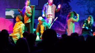 Энергичная массовая сцена: разноцветная подсветка, актёры в движении и плотная композиция на сцене с множеством жестов.