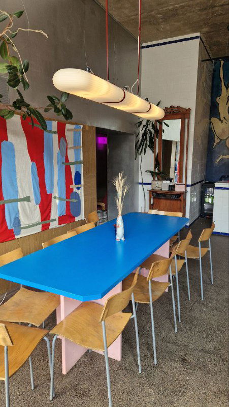 Длинный синий стол и стулья в светлой части бара, над столом висят современные подвесные лампы, на стене абстрактный арт‑объект.