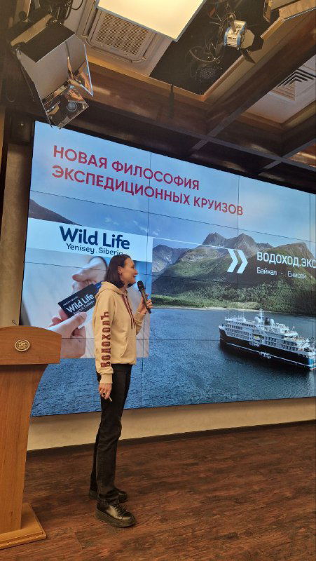 Спикер на сцене у большого экрана с заголовком «Новая философия экспедиционных круизов», кадр пресс‑завтрака в здании Русского географического общества.