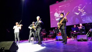 Исполнители в движении: скрипач, гитарист и басист на переднем плане, яркая боковая подсветка и затемнённый зрительный зал на заднем фоне.