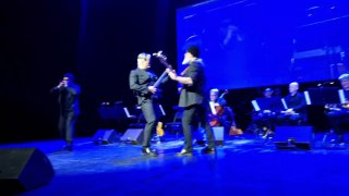 Размытый динамичный кадр с музыкантами на сцене: гитара и скрипки в игре, заметно сценическое освещение и движение артистов.