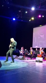 Вертикальный кадр: солист у переднего плана сцены, за ним — струнный ансамбль и проекционный экран, видно сценическое оформление.