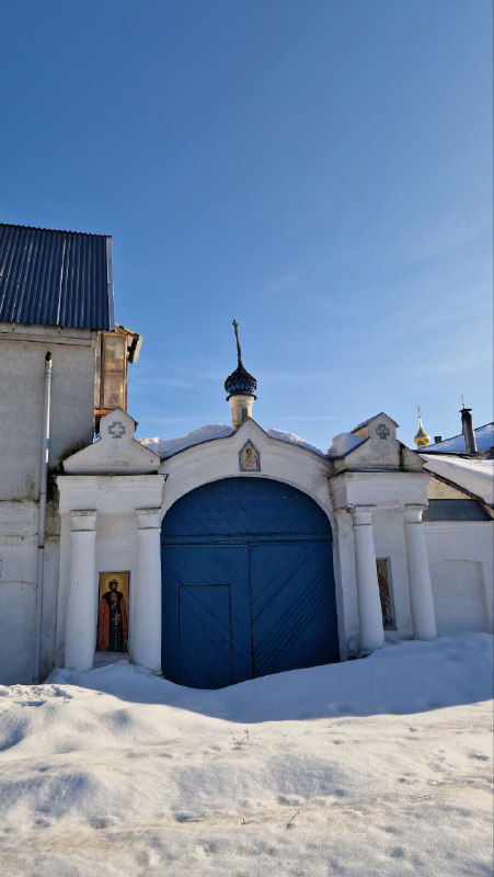 Ворота монастыря с массивными синими дверями и сугробами снега на переднем плане, ограждение и хозяйственные постройки на заднем плане.