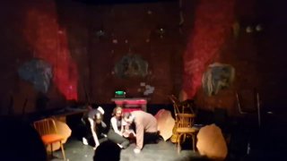 Групповая сцена на небольшой сцене: актёры собраны вокруг стульев, приглушённое освещение и проекции на стенах создают напряжение.