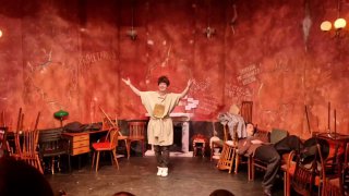 Актор в костюме на фоне красноватых стен и расставленных стульев; яркое световое решение подчёркивает театральную экспрессию.