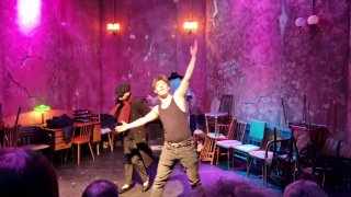 Динамичная сцена с двумя актёрами в движении, разноцветное световое решение и множество стульев как элемент сценографии.