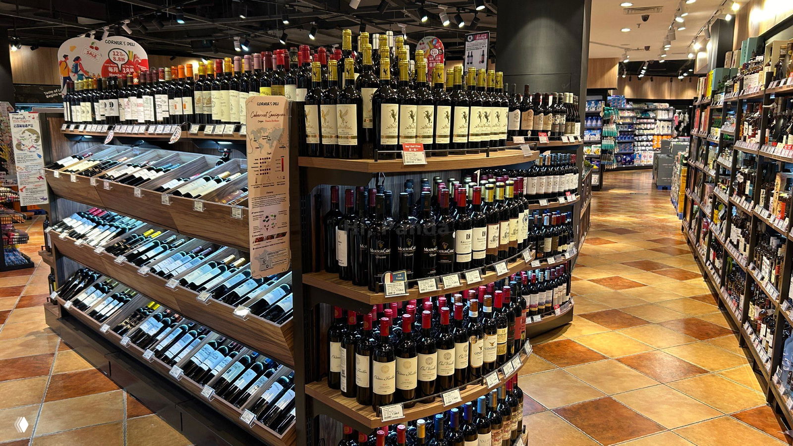 Круглая демонстрационная стойка и наклонные лотки с бутылками красного и белого вина, освещённый проход в супермаркете внутри ТЦ «Лето».