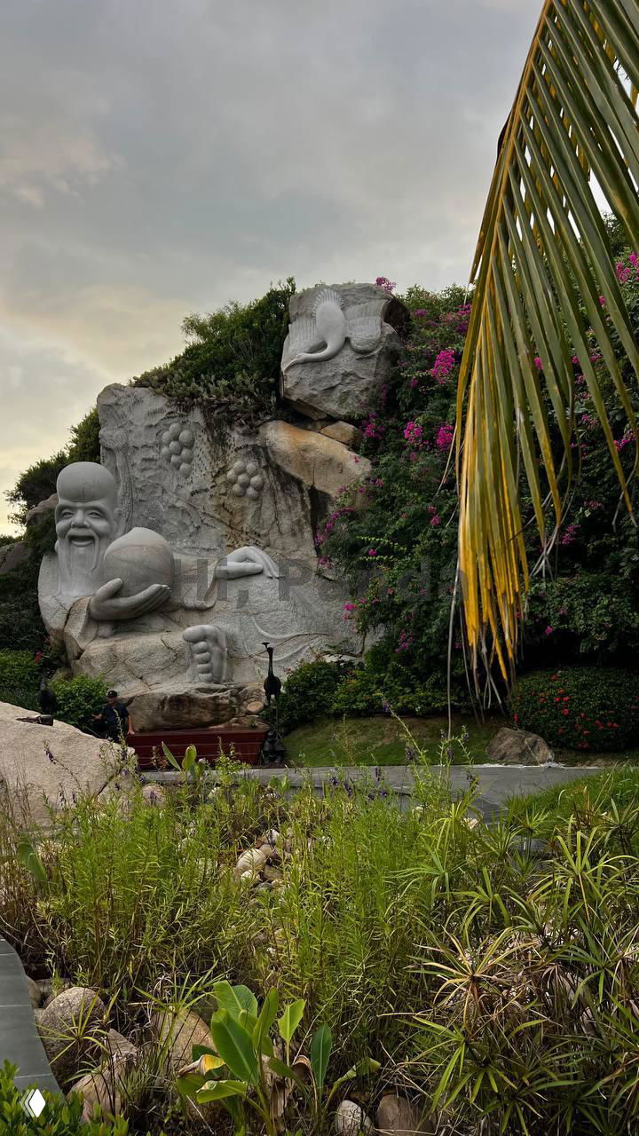 Каменная скульптура мудреца с шаром в руках в зелёной прибрежной зоне парка, заросли, тропинки и неяркий вечерний свет.