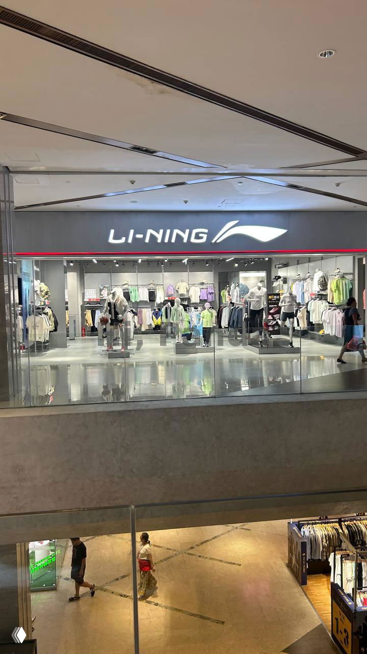 Светящаяся вывеска Li-Ning на втором этаже: спортивный магазин с манекенами и одеждой, вид через перила галереи торгового центра.