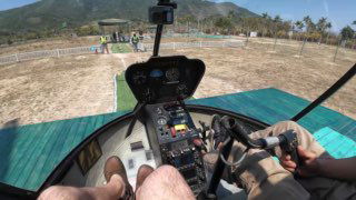 Вид из кабины вертолёта на приборную панель, ноги пилота и посадочную площадку на фоне гор и побережья на Хайнане.