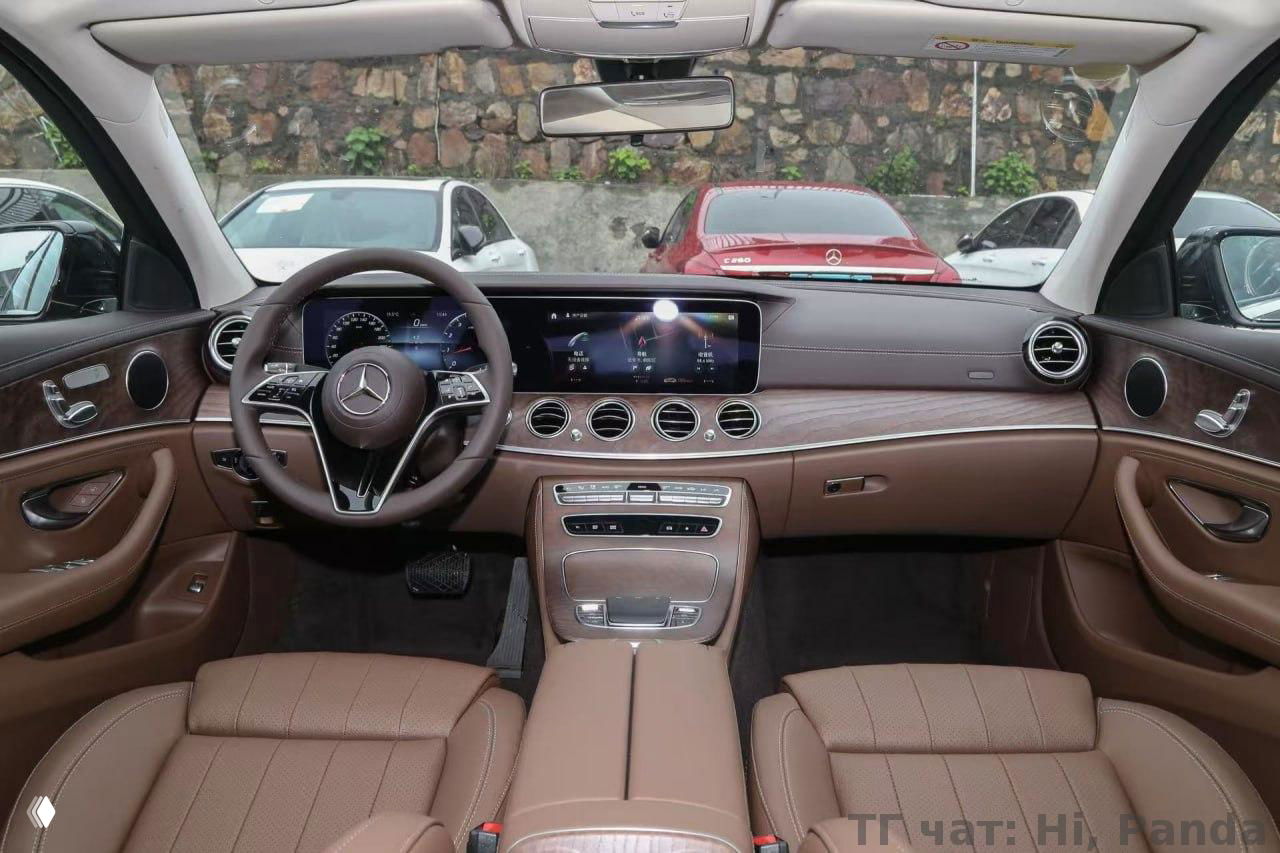 Интерьер Mercedes: тёплая коричневая кожа сидений, широкая панель с большим экраном и элементы отделки под дерево, вид из переднего ряда салона.