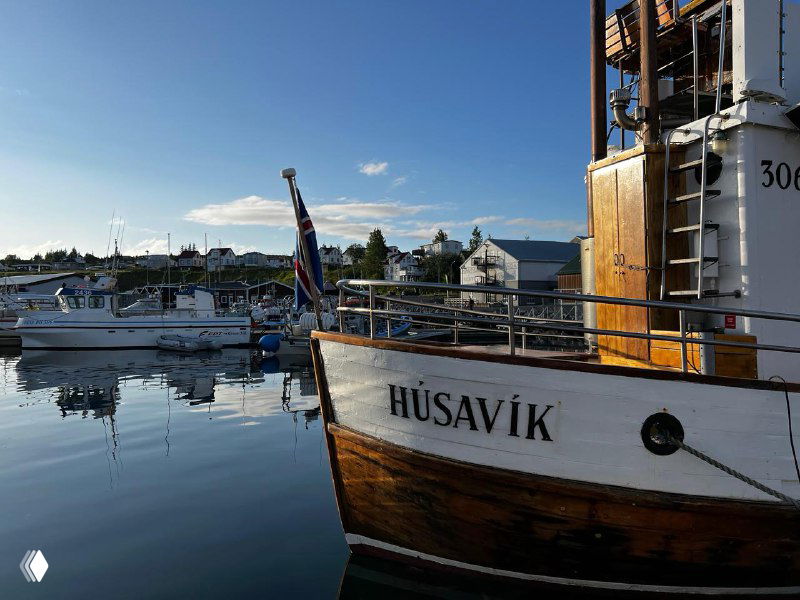 Деревянный прогулочный корабль с надписью HÚSAVÍK пришвартован в тихой гавани; на заднем плане причалы и здания порта.