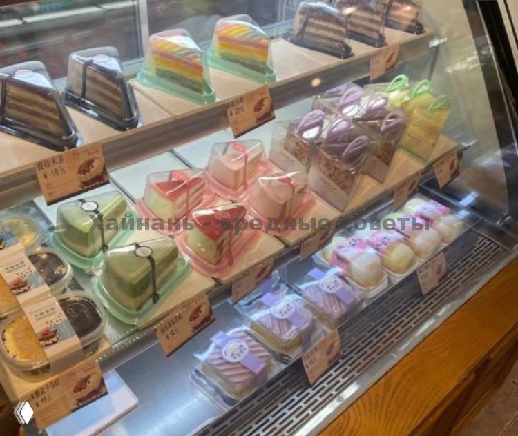 Холодильная витрина с порционными тортами и десертами в коробках, аккуратная расстановка, ценники и яркая подсветка, интерьер кондитерской на Хайнане.