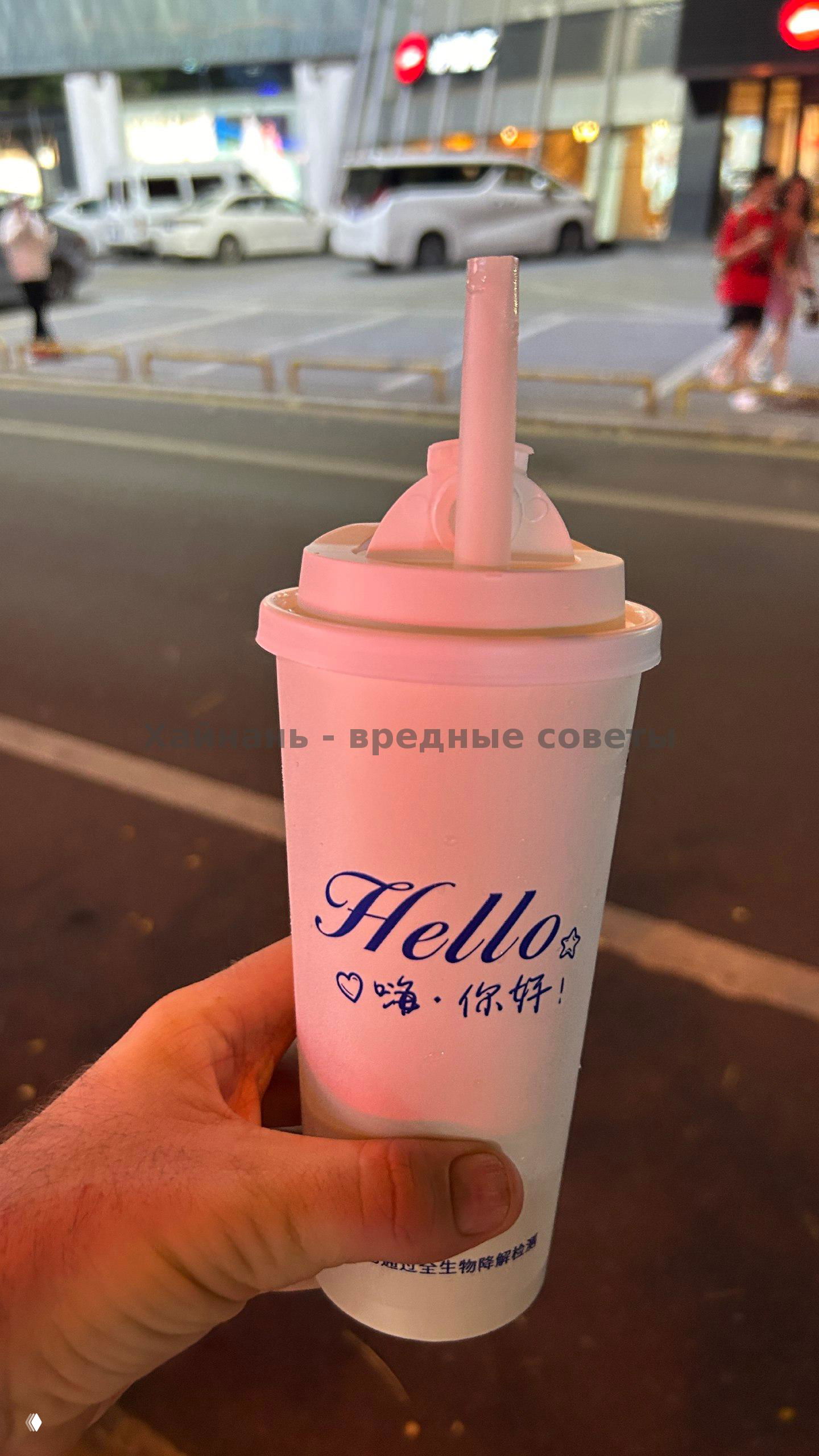 Рука держит высокий бумажный стакан с крышкой и надписью «Hello», на фоне вечерняя улица и припаркованные машины — напиток с собой на улице в Хайнане.