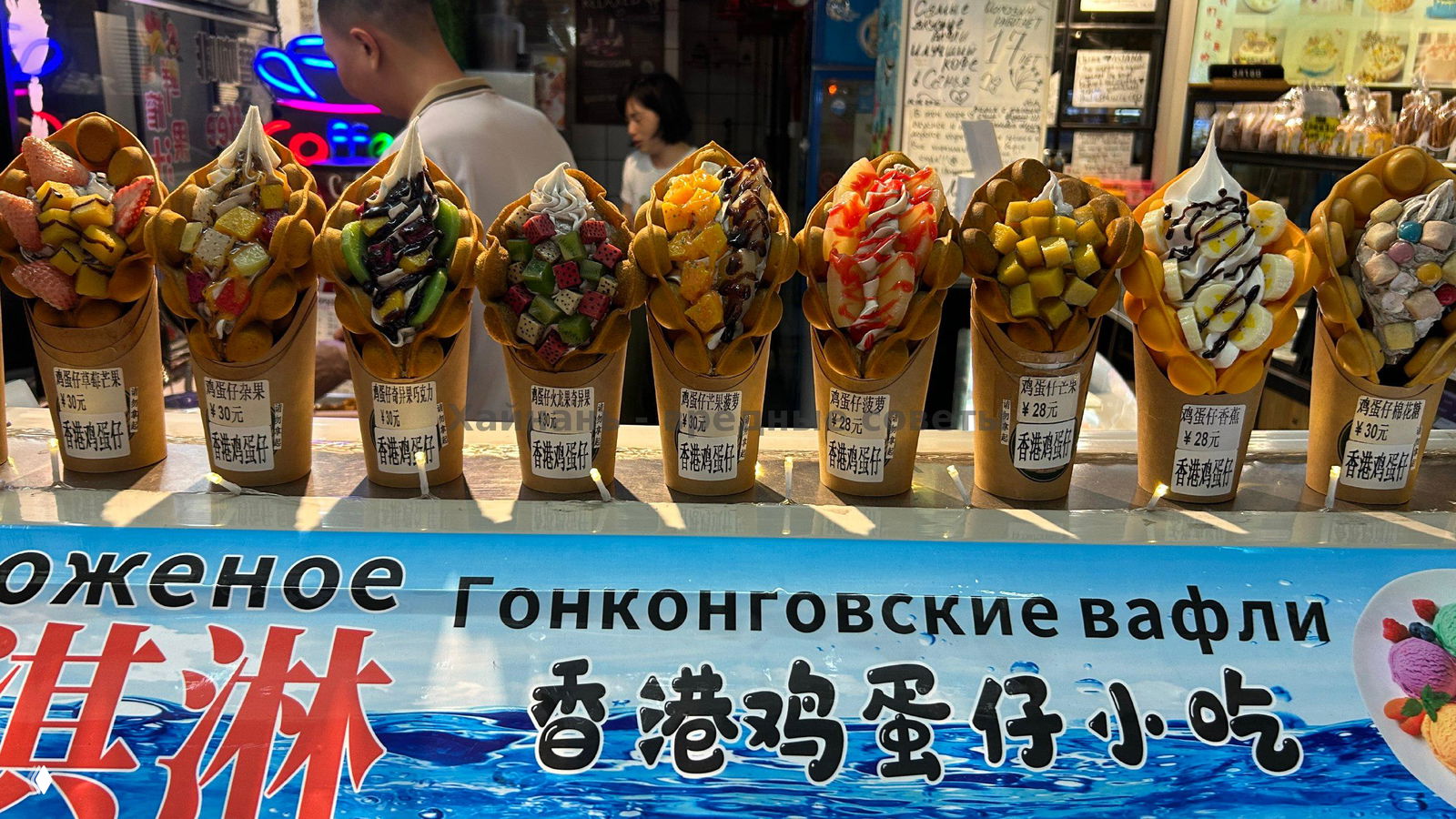 Панорамный вид прилавка с множеством вафель на фоне баннера «Гонконгские вафли», русские и китайские надписи, освещение прилавка вечером на улице Хайнань.