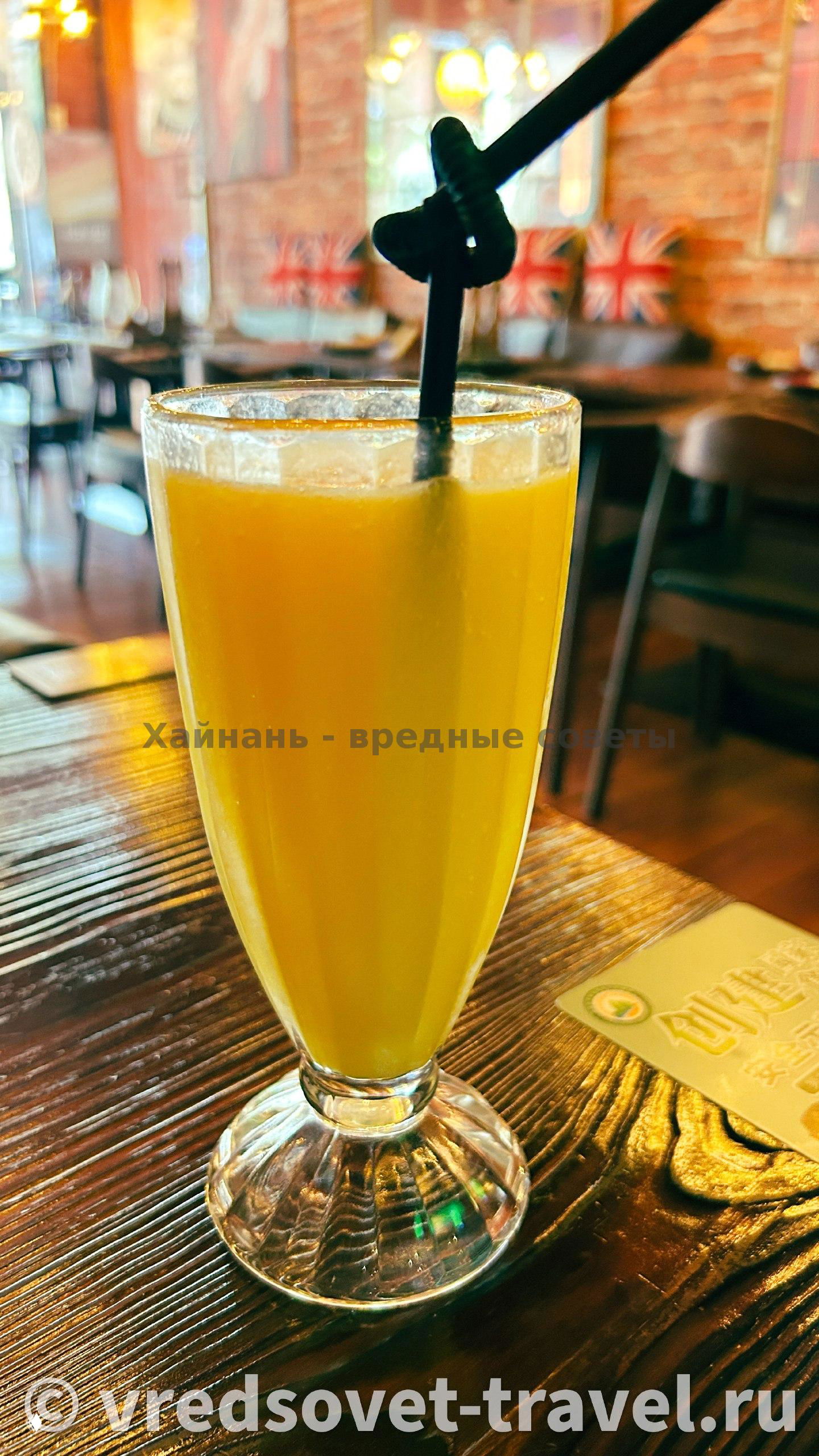 Высокий бокал апельсинового сока на столе внутри ресторана, фон с креслами и украшениями в европейском стиле, логотип сайта на фото.