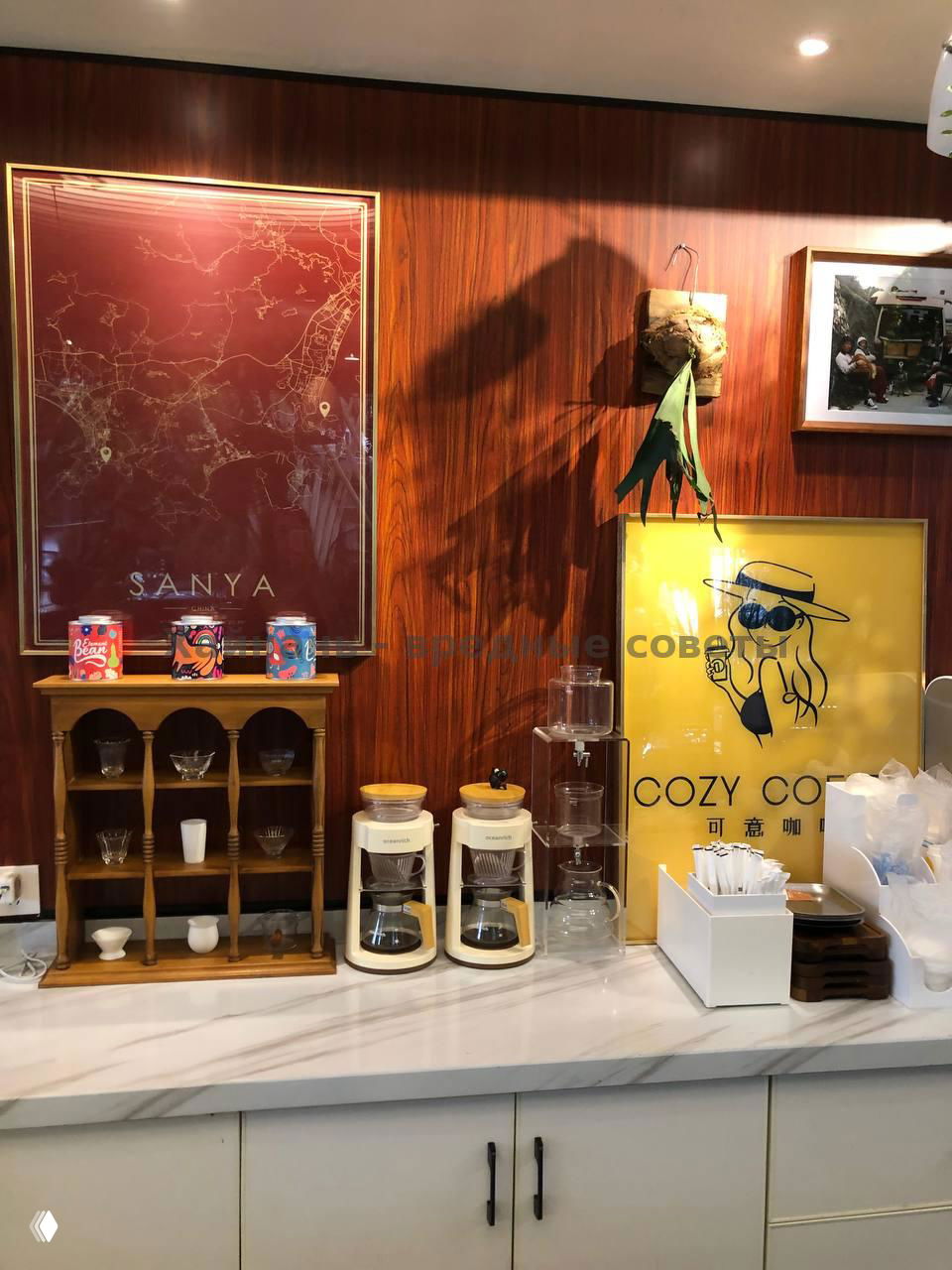 Прилавок и оборудование: карта Саньи на стене, кофейные приборы и два капельных кофеварочных аппарата на белой столешнице в интерьере Cozy Coffee.