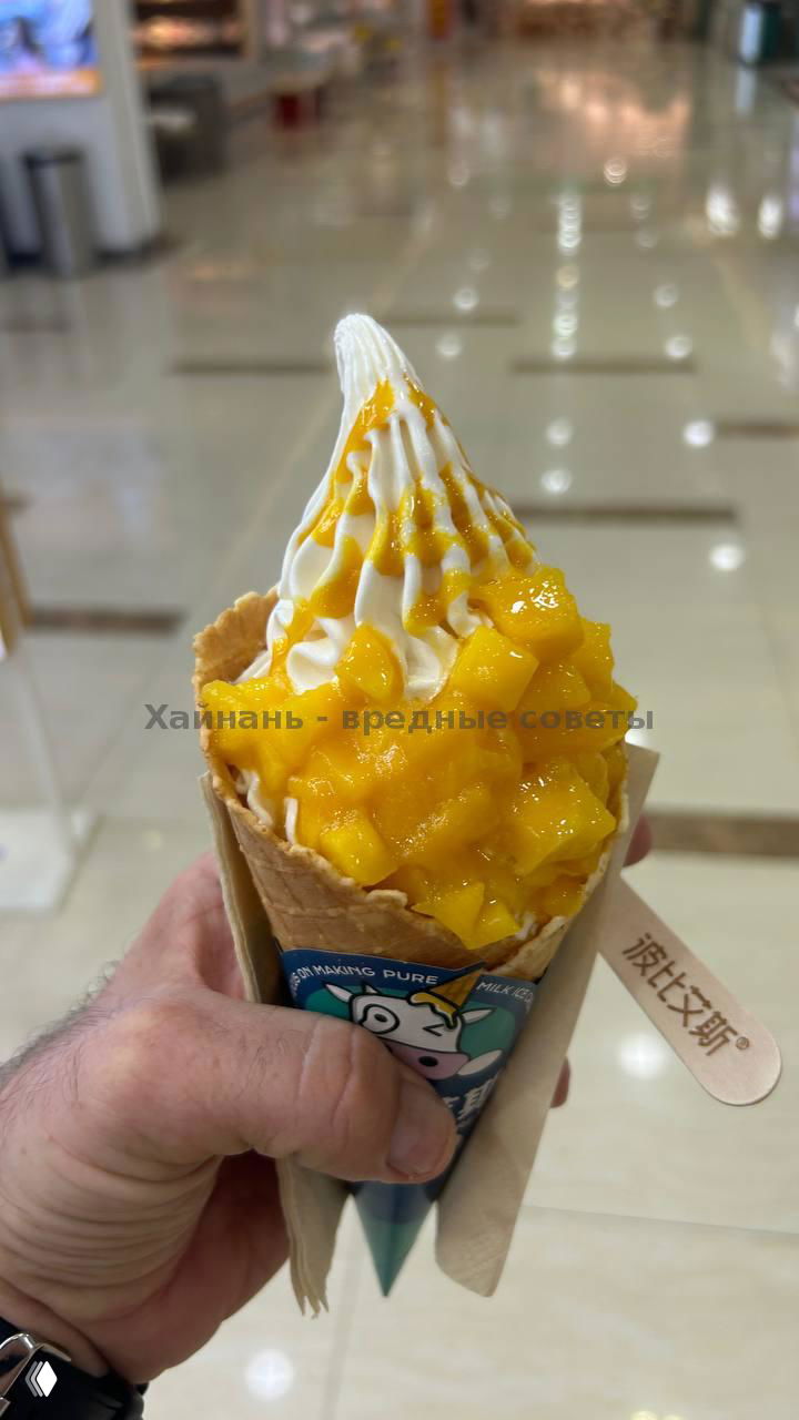 Рожок с мягким мороженым и крупно нарезанными кусочками манго сверху; держат в руке, фон — размытый интерьер торгового центра.