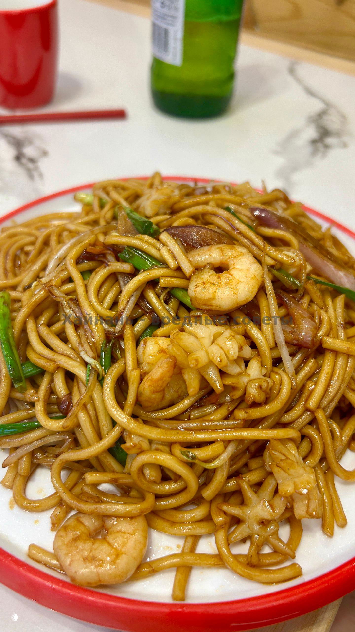 Крупный план жареной лапши с креветками и овощами: золотистые нудлы, зелёный лук и кусочки морепродуктов на белой тарелке с красной каймой.