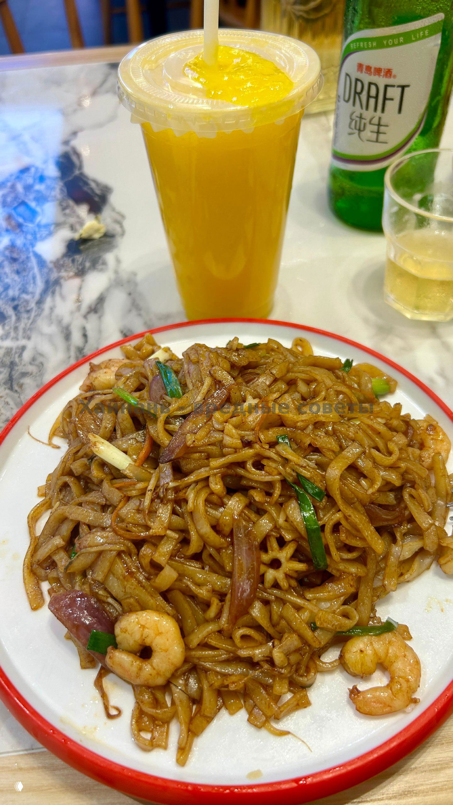 Стакан мангового сока и тарелка жареной лапши на столе ресторана; на заднем плане бутылка пива и столовая посуда, кадр показывает напиток и блюдо вместе.