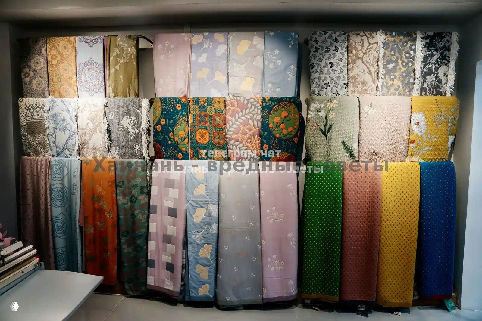 Стойка с рулонами декоративных тканей и материалов: яркие узоры и текстуры, представленные вертикально на стенде магазина.