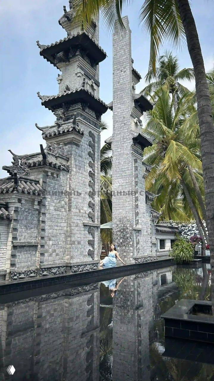 Каменные пагоды и башни буддистского комплекса Наншань среди пальм, с зеркальным бассейном внизу и туристкой у воды.