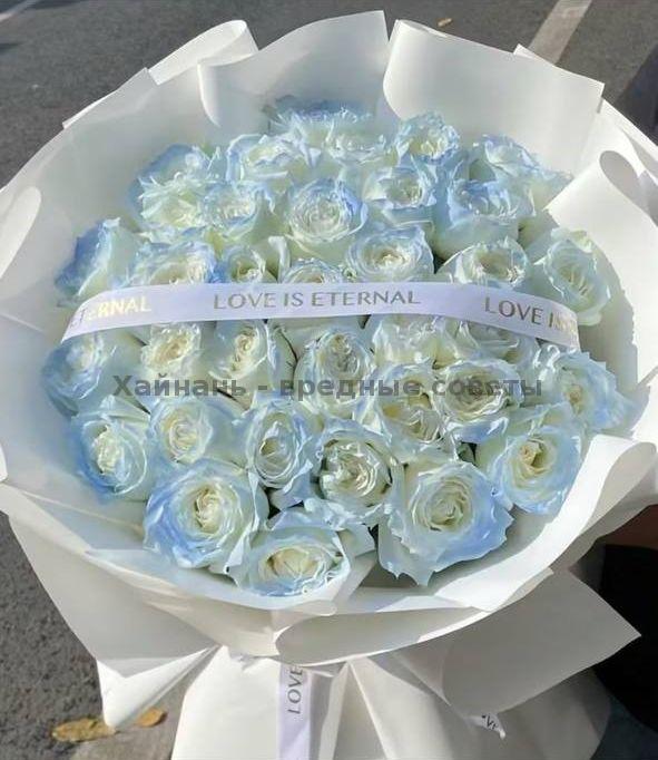 Большой монобукет белых роз с лёгким голубоватым оттенком, оформлен в белую матовую бумагу с лентой «LOVE IS ETERNAL», фото подчёркивает объём и текстуру цветов.