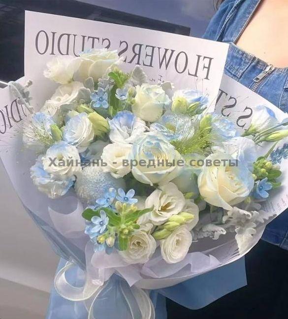 Пышный букет бело-голубых и кремовых цветов с зелёными акцентами в фирменной обёртке с логотипом FLOWERS STUDIO, держат на руках на фоне одежды.
