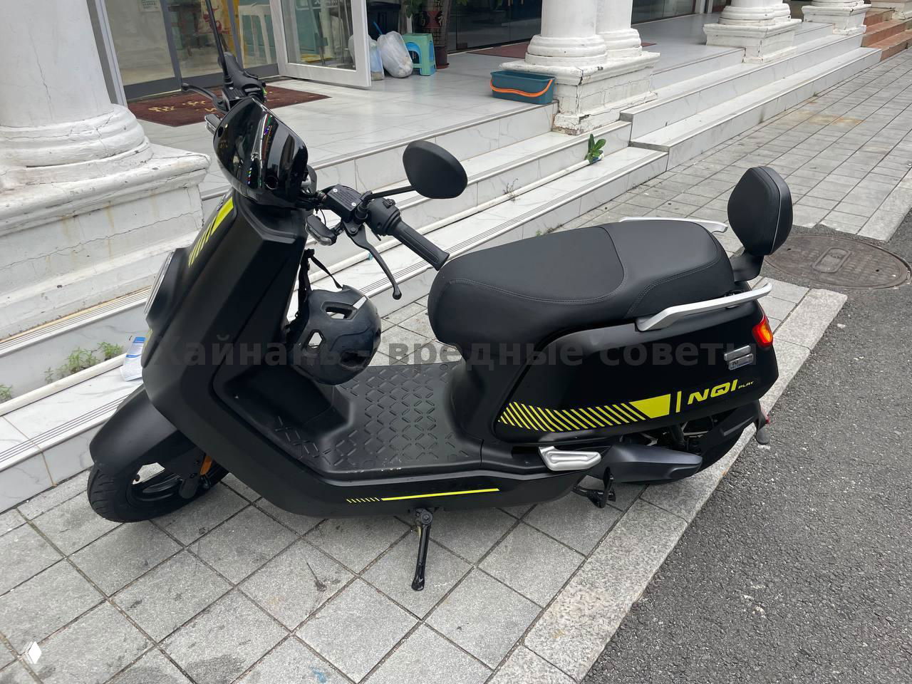 Ещё один боковой вид чёрного электрического скутера NIU на плиточном покрытии у входа в здание, видна сидушка, поручень и защитный шлем в полке.