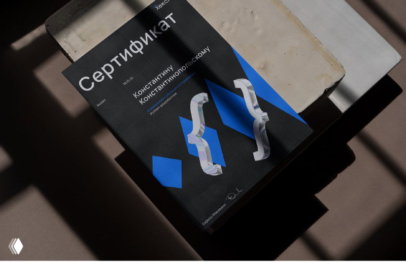 Фотография сертификата Hexlet на столе: тёмный дизайн с синими ромбами и геометрическими акцентами для брендированной печатной продукции.