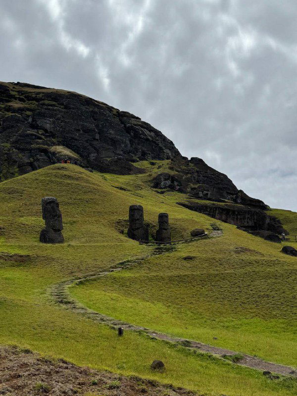 Холмы острова Пасхи с расставленными по склону частично видимыми головами моаи, тропинка ведёт вдоль зелёного рельефа под облачным небом.
