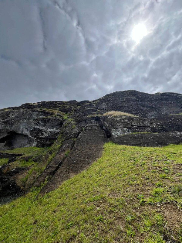 Крутой скалистый склон и выветренная поверхность вулканической горы на острове Пасхи под напряжённым облачным небом, вид снизу вверх.