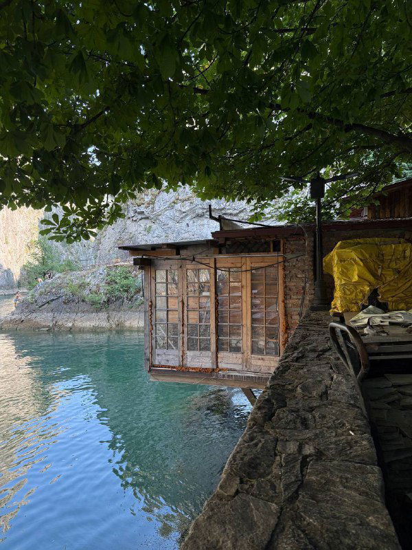 Небольшая деревянная пристань и домик у скалы под навесом, спокойная вода и тень от листвы в природном резервате.