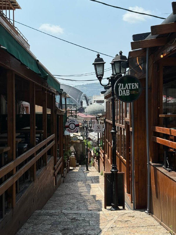 Узкая пешеходная улица в старом городе с деревянными лавками и вывесками, туристическая атмосфера и вид на холмы.