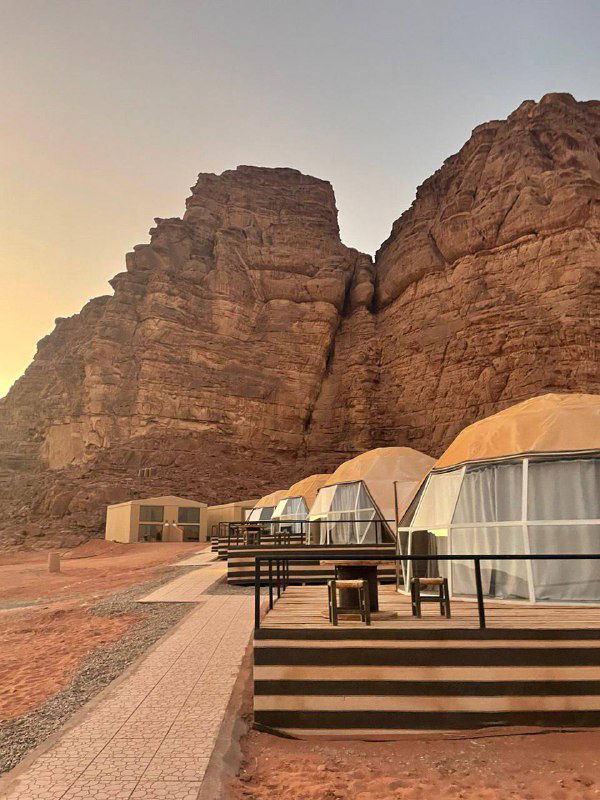 Глэмпинг-купола и деревянные террасы у подножия внушительных скал в пустынном ландшафте, вечерний свет и спокойная обстановка.
