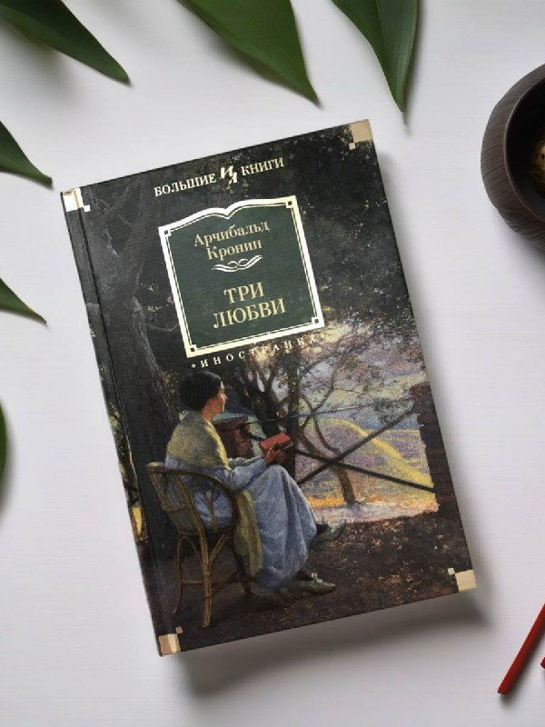Книга «Три любви» Кронин: обложка с иллюстрацией; издание лежит на столе рядом с чашкой и зелёными листьями в уютной композиции.