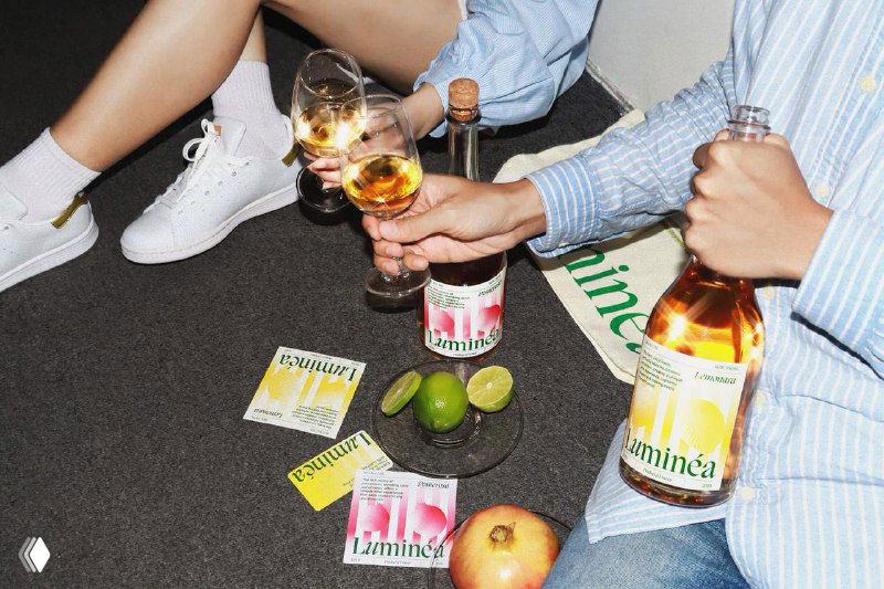 Молодёжная сцена с людьми, держащими бутылки и бокалы Luminea Wine; рассыпанные упаковки и фрукты подчёркивают неформальную подачу бренда.