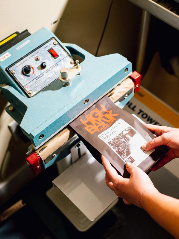 Процесс упаковки: руки оператора работают с запаивателем и приклеивают или прогревают большую бумажную этикетку High Bank на пакет.