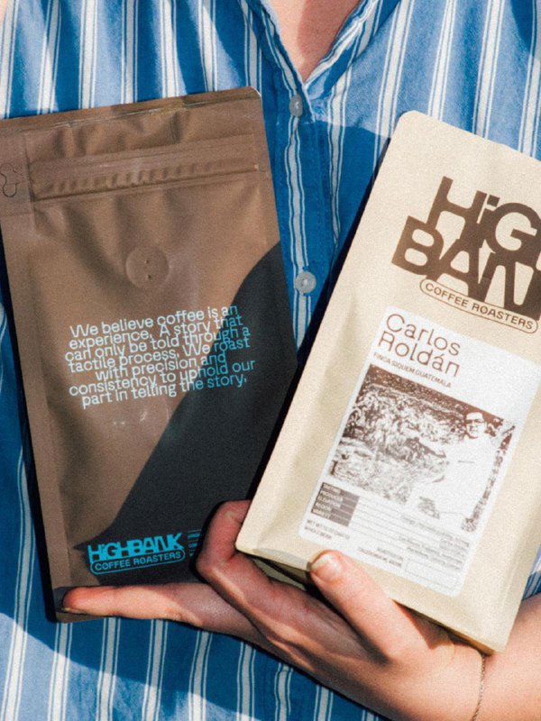 Крупный план: человек держит две упаковки High Bank, на белой этикетке видно имя фермера и чёрно‑белую фотографию производителя зёрен.