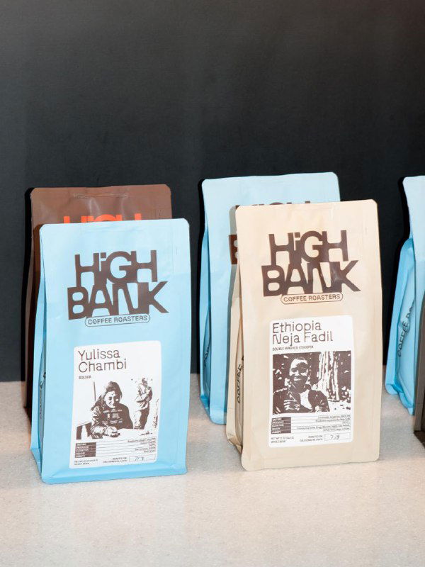 Ряд разноцветных упаковок кофе High Bank на столе; у каждой большая прямоугольная этикетка с фото фермера и информацией о регионе происхождения.