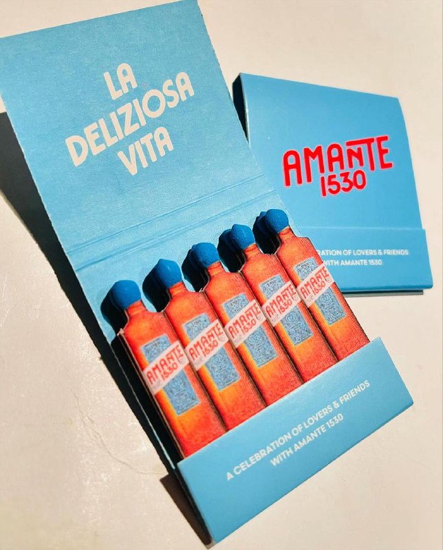 Голубая коробка спичек с иллюстрацией бутылок и надписью La Deliziosa Vita: яркие оранжево‑синие наконечники, композиция в винтажном ключе, стилизованная под продуктовую рекламу.